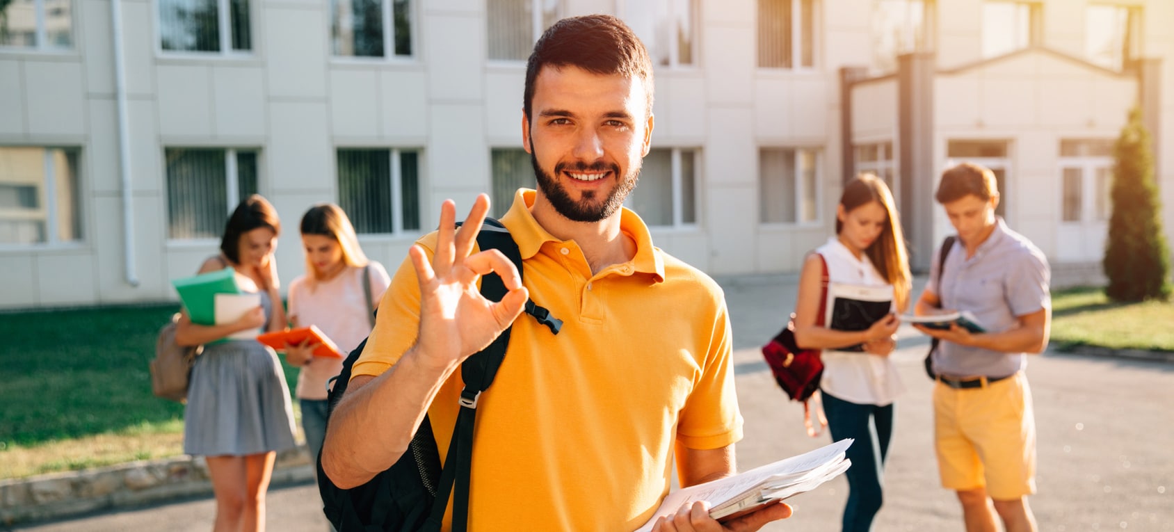 Happy student with note-books and backpack smiling and showing okay while standing on background of university and friends